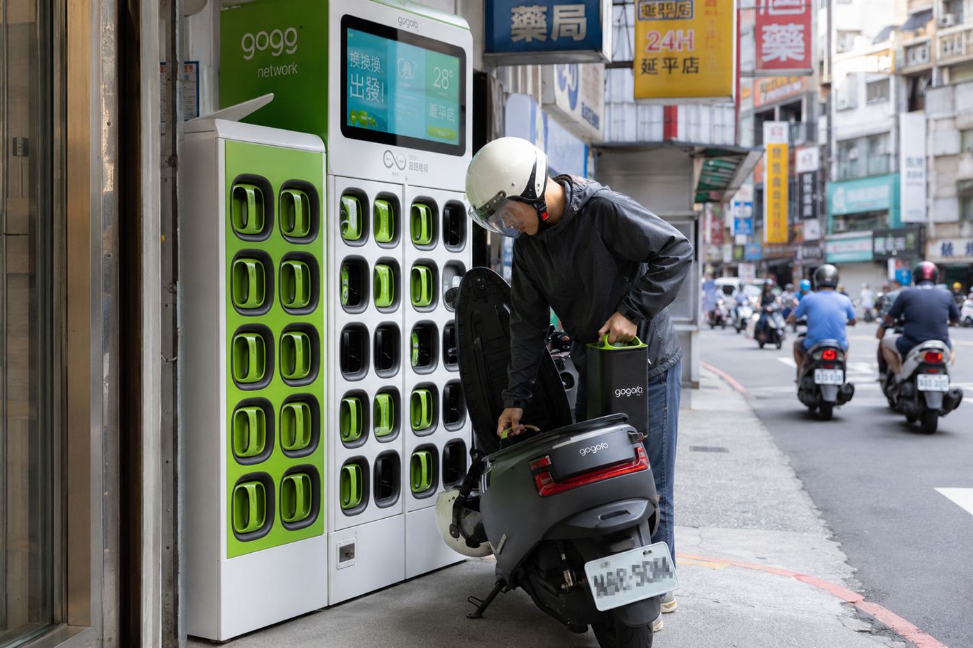 A rider at a bright green Gogoro GoStation kiosk on a Taiwan street, lifting a battery out of an electric scooter to swap it