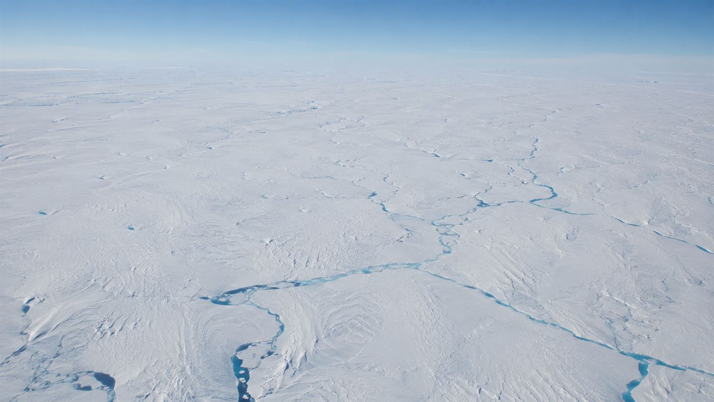 An aerial view of the vast white Greenland ice sheet stretching to the horizon with faint blue meltwater