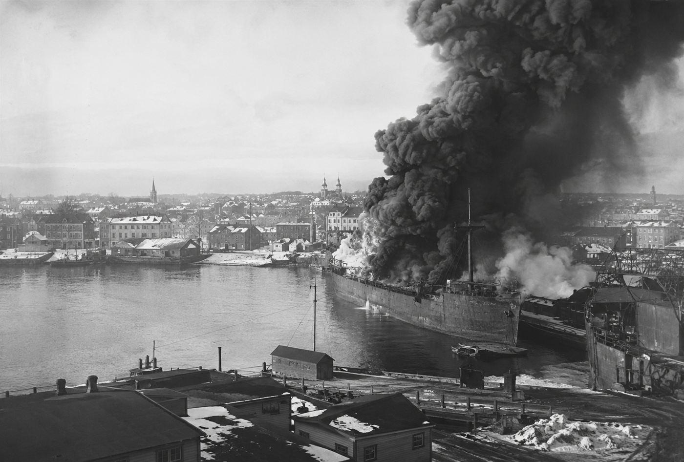 A large munitions ship on fire in Halifax Harbour sending thick black smoke into the winter sky of 1917, Canadian port buildings visible on the waterfront, dramatic documentary historical scene