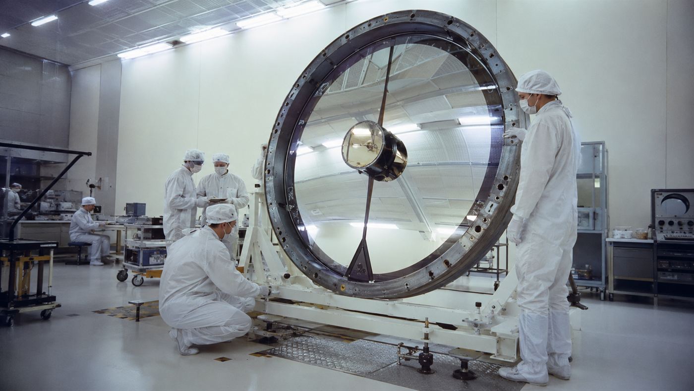 Engineers in a clean room inspecting the large circular primary mirror of a space telescope