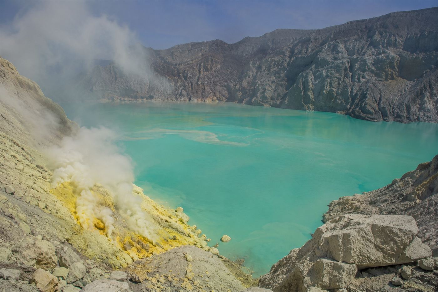 The turquoise acidic crater lake of Kawah Ijen in daylight with yellow sulfur deposits on the shore