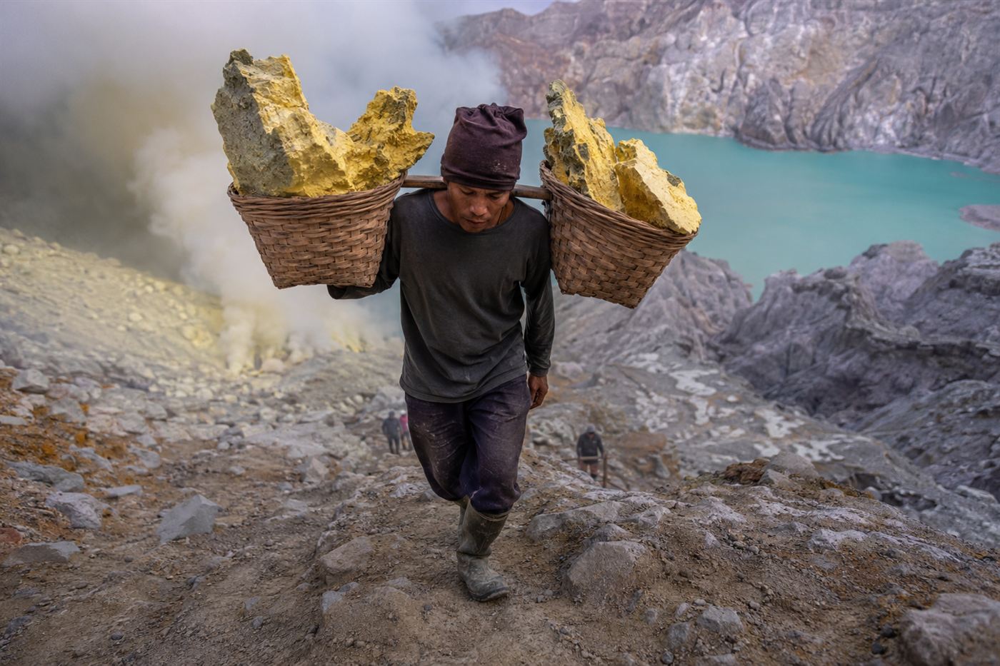 A sulfur miner carrying two heavy baskets of yellow sulfur on a pole up the steep path out of the Kawah Ijen crater