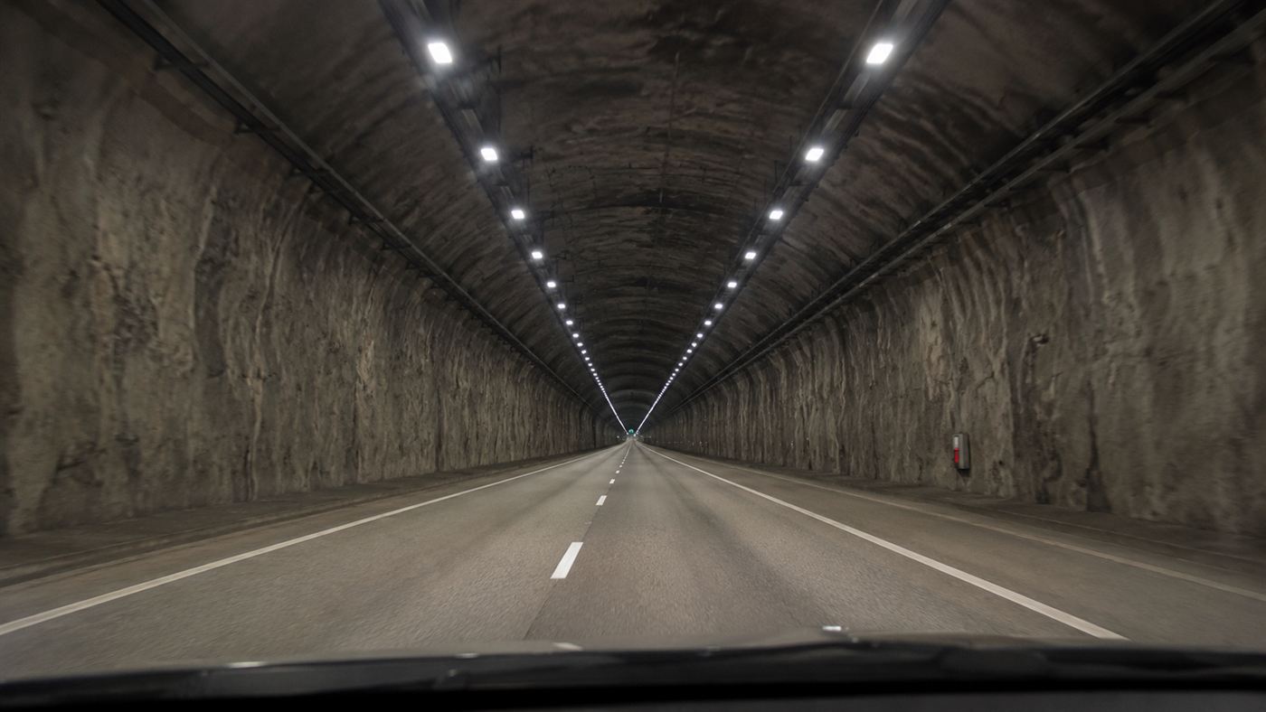 A car's view down a long straight rock tunnel with rows of lights stretching far into the distance