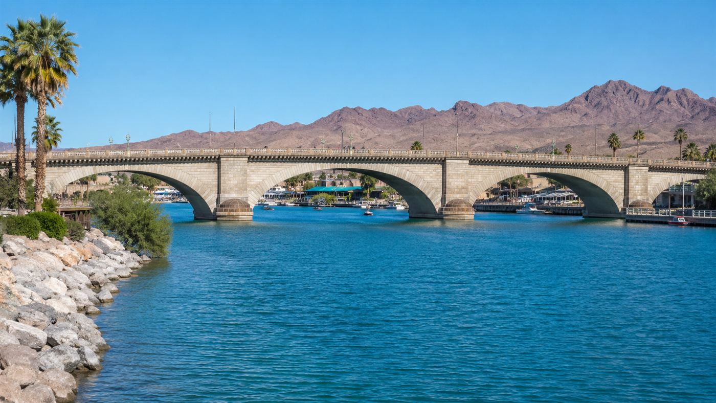 London Bridge, a long pale granite arch bridge, spanning a blue channel in the Arizona desert with rocky hills and palm trees under a bright sky