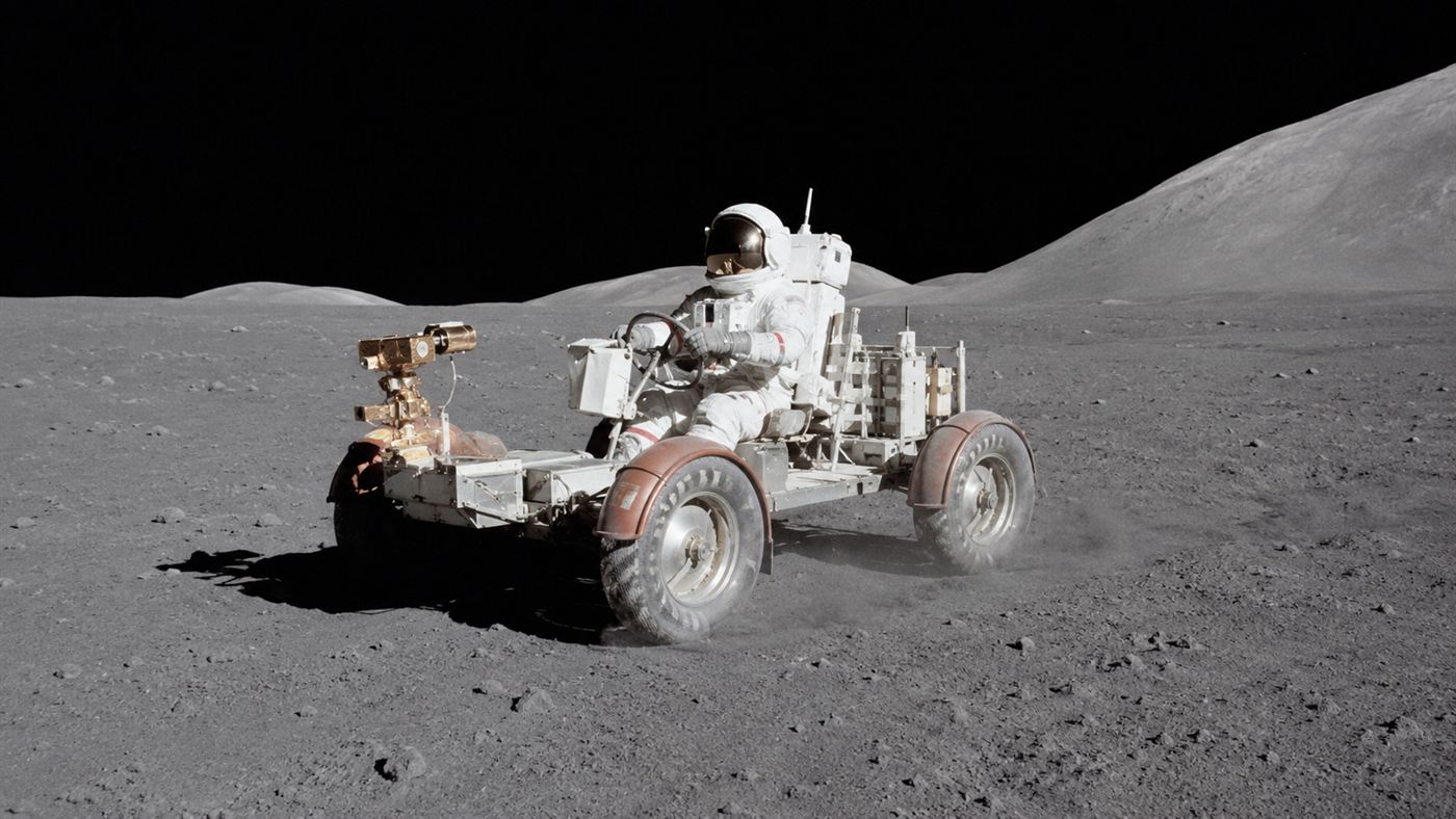An astronaut driving the Apollo Lunar Rover across the grey dusty surface of the Moon under a black sky