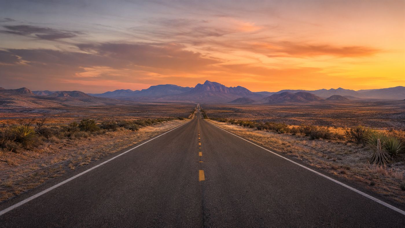 An empty straight desert highway stretching toward distant mountains in west Texas at dusk, near the Marfa lights viewing platform