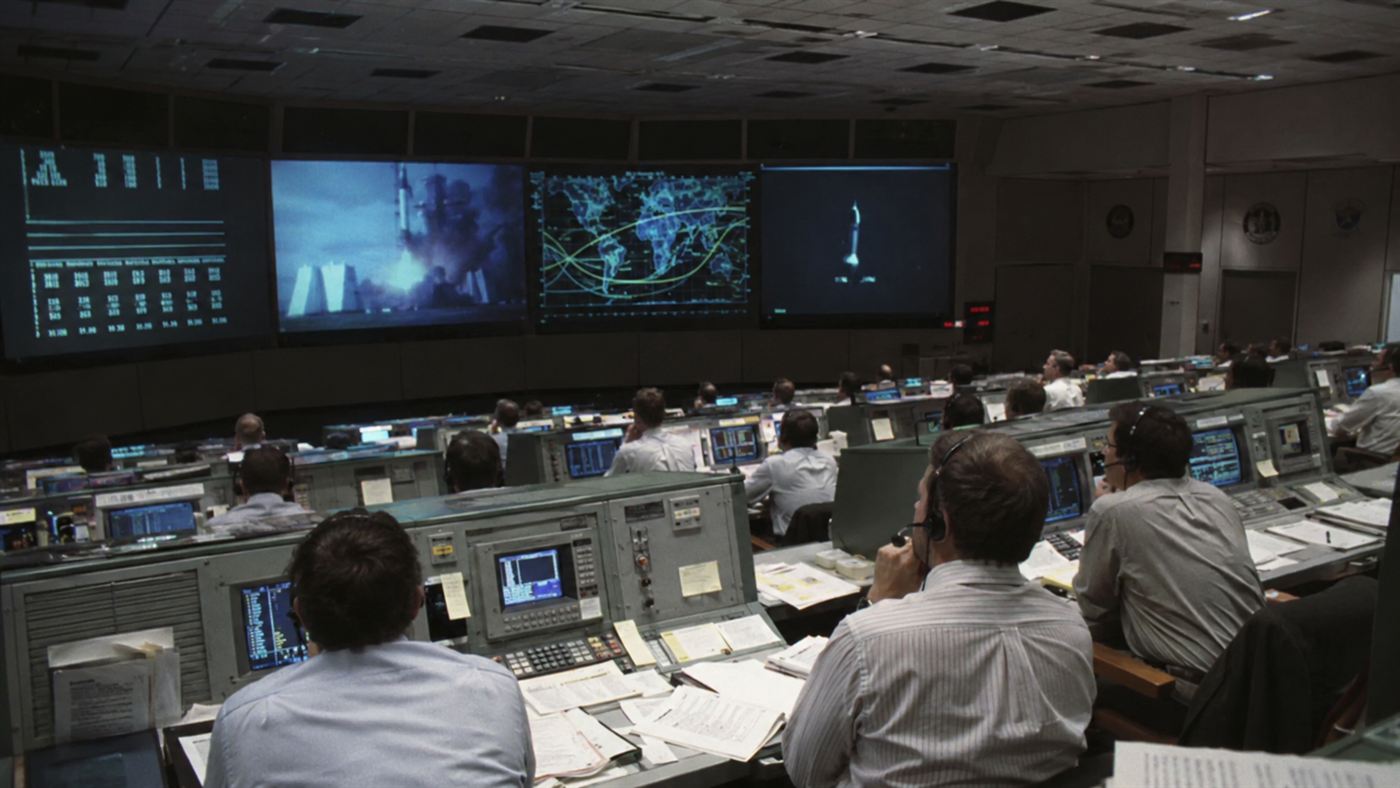 A 1990s NASA mission control room with rows of engineers at consoles watching large screens