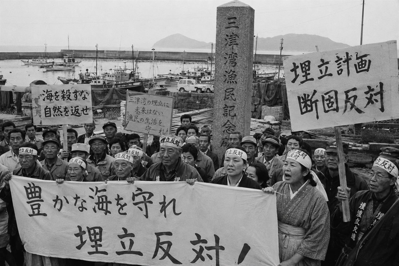 Japanese Minamata disease victims and their families carrying protest banners near a stone waterfront memorial, demanding justice and recognition from Chisso Corporation in the 1970s, with fishing boats visible in the harbor behind them