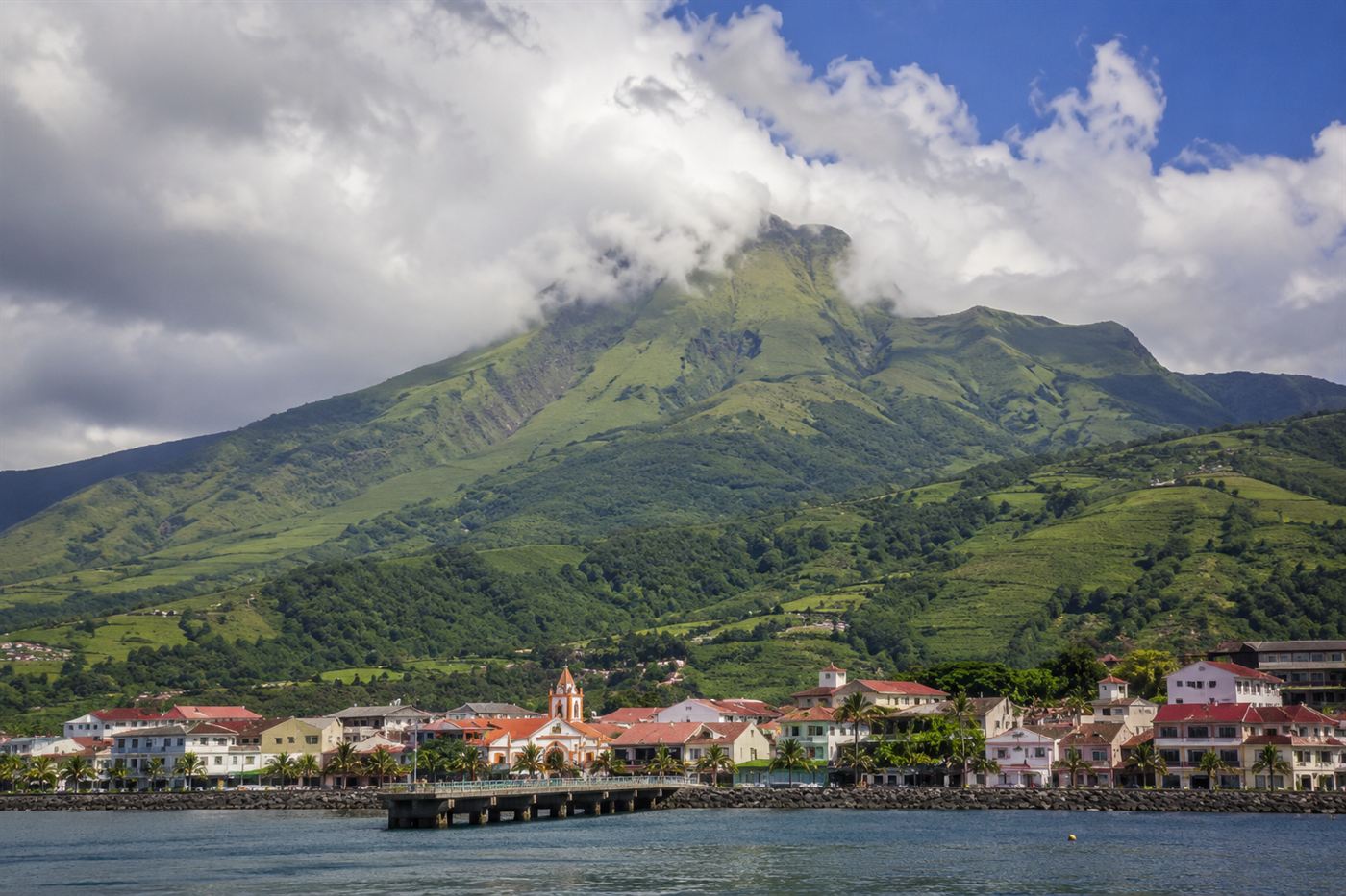 The lush green volcanic peak of Mount Pelée rising above the coastal city of Saint-Pierre in Martinique, Caribbean tropical landscape with dramatic clouds around the volcano summit