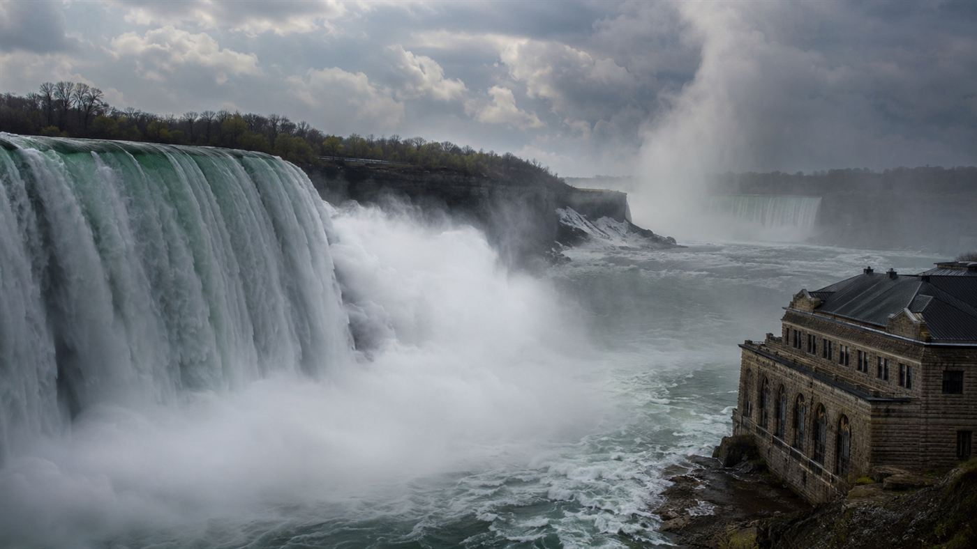 Niagara Falls thundering over the cliff in clouds of mist with an old stone hydroelectric powerhouse on the riverbank