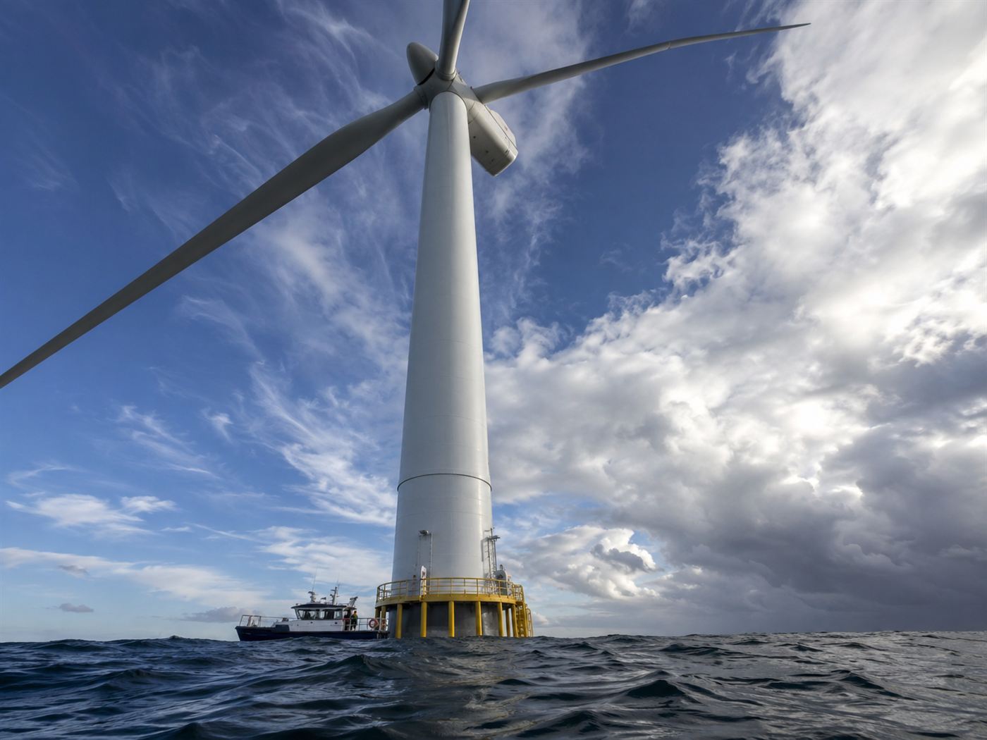 A crew boat dwarfed beneath the enormous tower and blades of an offshore wind turbine, showing the scale of the machine