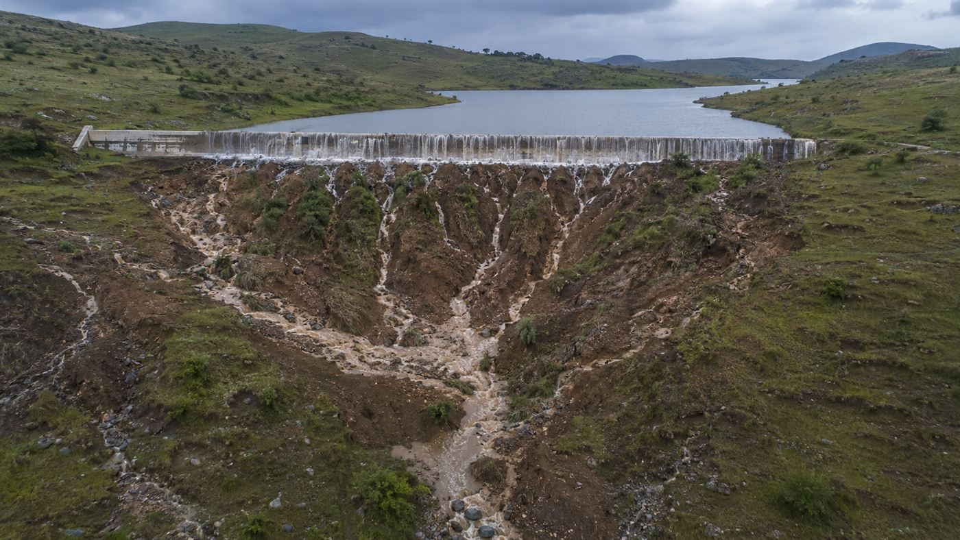 Water spilling over a long concrete emergency weir with deep raw erosion gullies cut into the bare hillside below, a spillway failure in progress