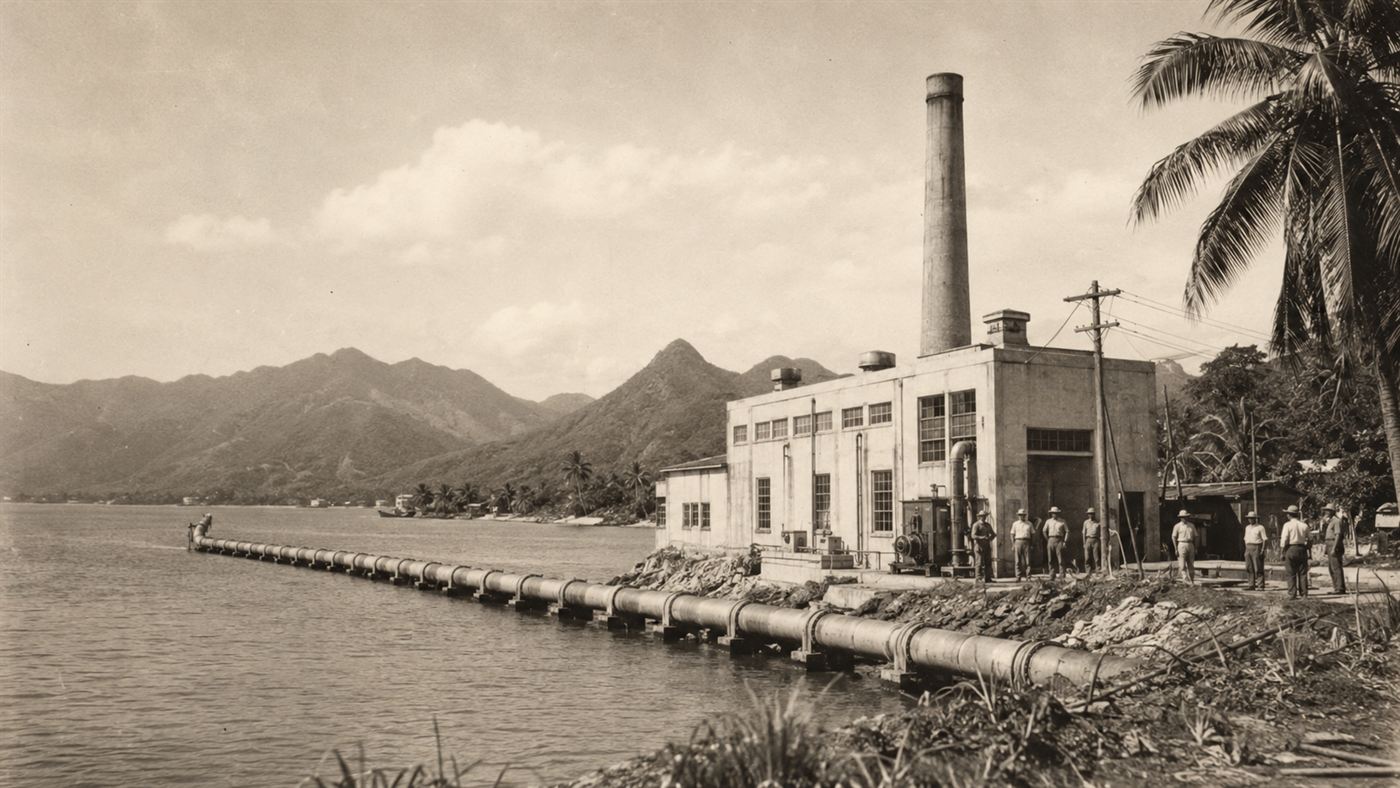 A 1930s shoreline experimental power plant with workers and a long pipe running out into a tropical bay