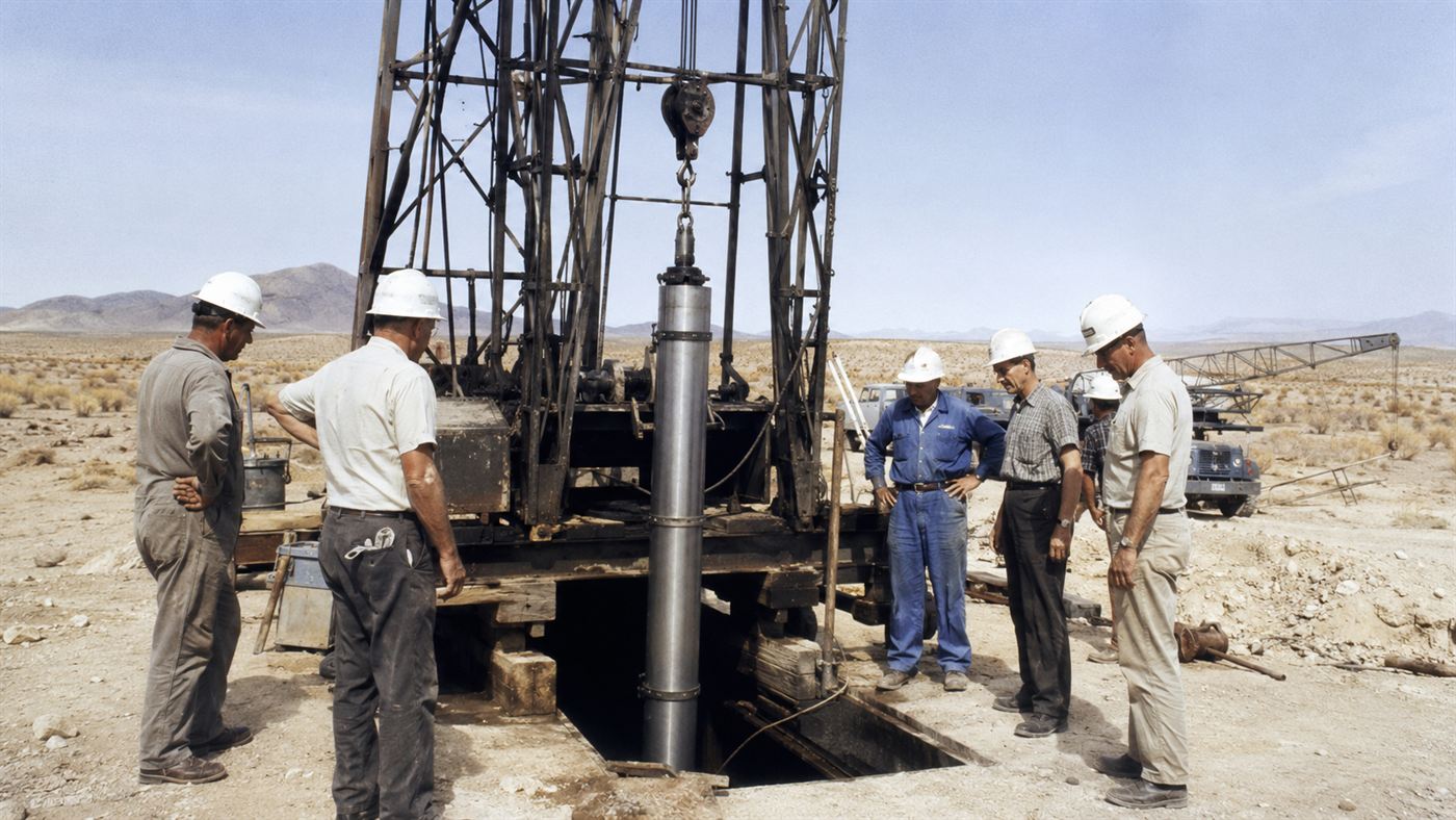 A 1960s deep borehole rig lowering a long cylindrical nuclear device down a shaft for an underground Plowshare test
