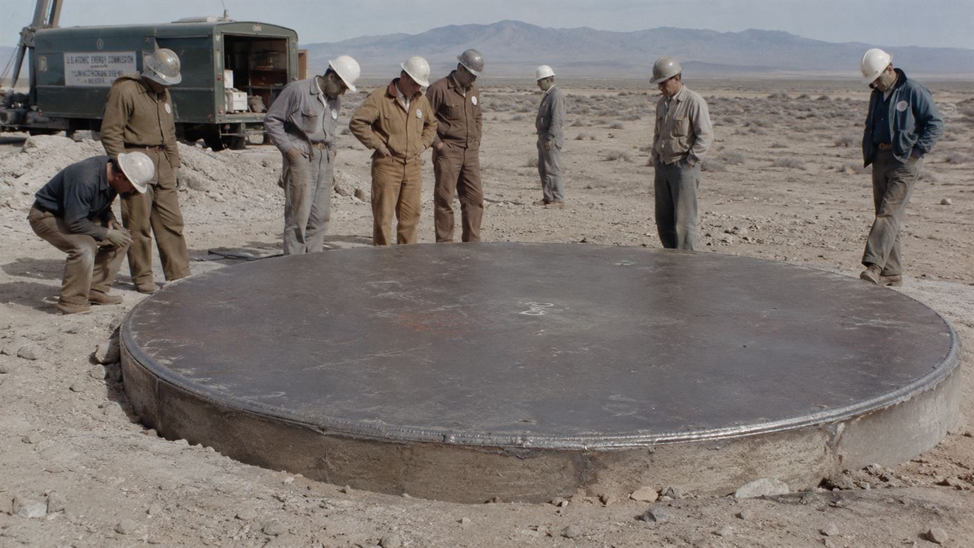 A heavy circular steel plate welded over a wide borehole in the desert, with 1950s workers nearby