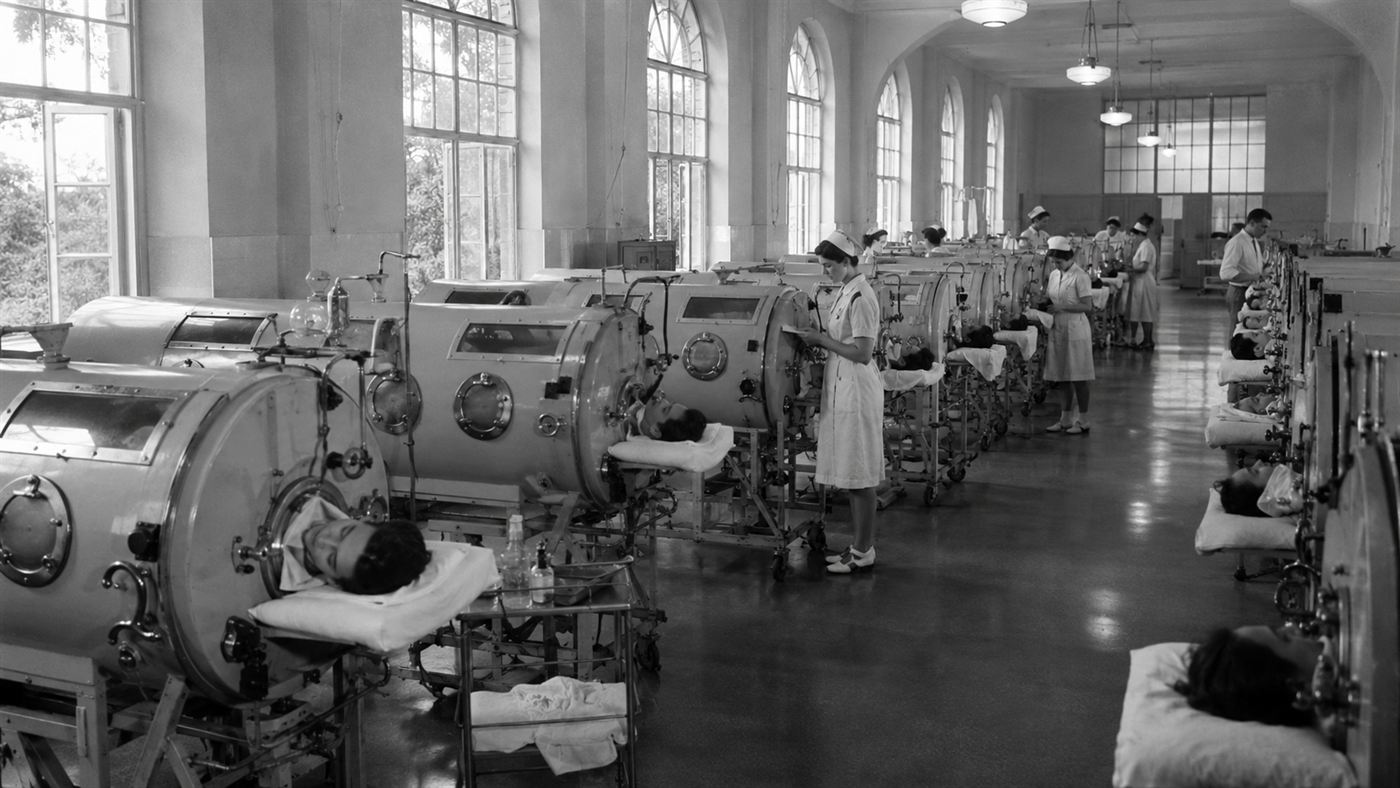 A 1950s hospital ward lined with rows of large cylindrical iron lung machines under tall windows
