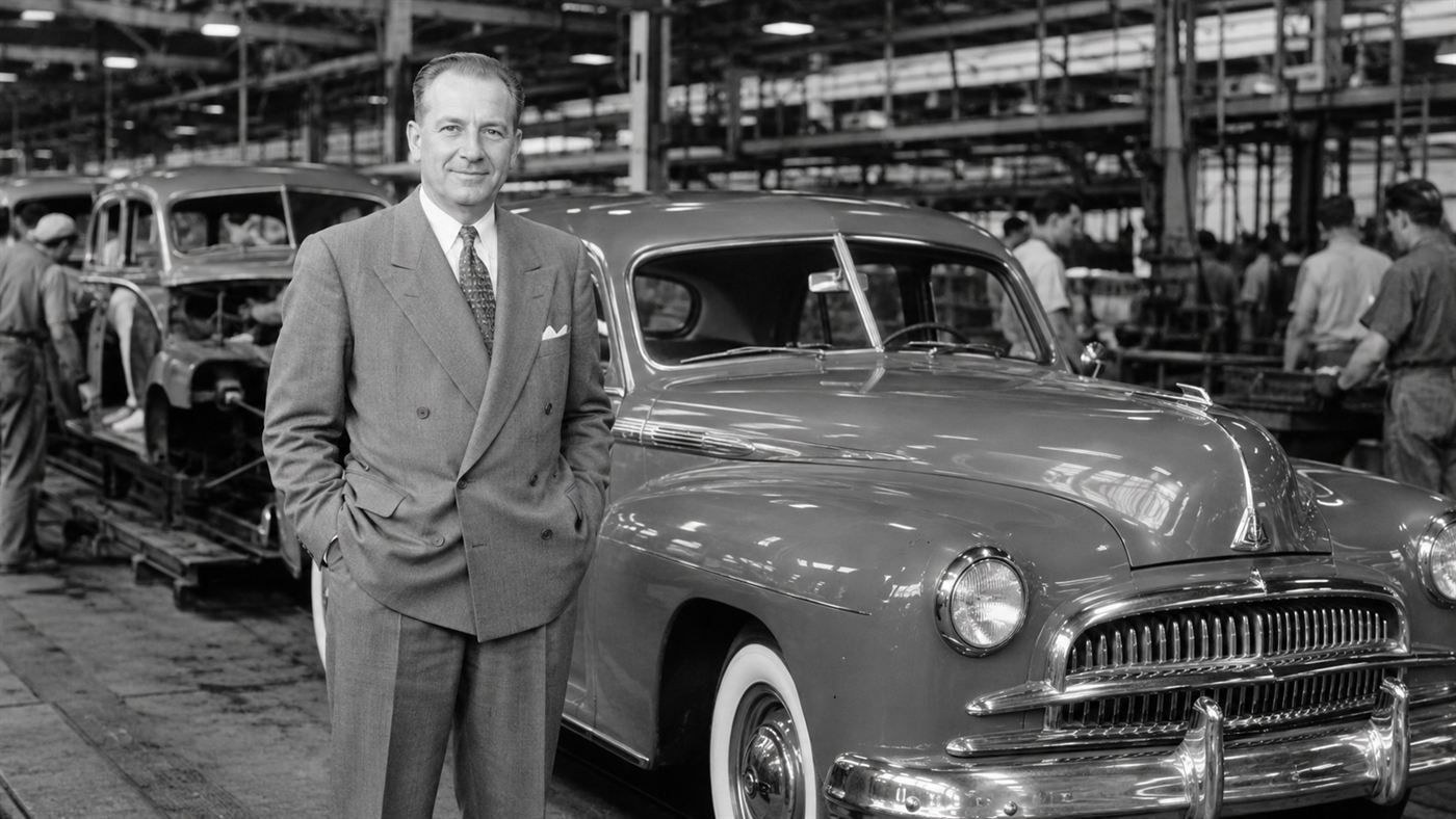 A 1940s businessman in a double-breasted suit standing beside a new sedan in a car factory