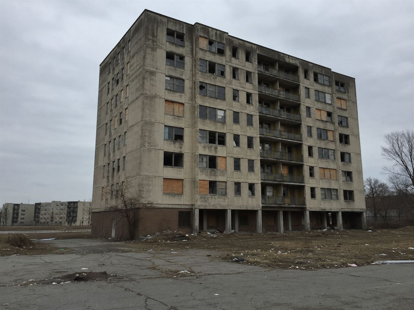 A weathered 1960s concrete public housing tower with many broken and boarded windows and litter on the empty grounds