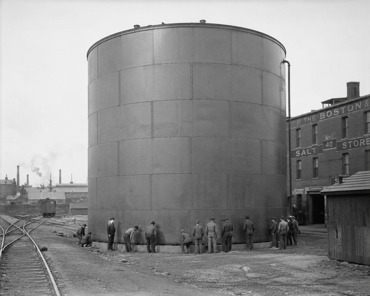 A large cylindrical steel industrial storage tank beside a railroad yard in an American industrial port city in 1915, workers examining the base, brick warehouse buildings in background