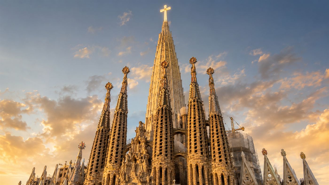 The Sagrada Familia basilica in Barcelona seen from below, its tall central tower crowned with a large cross rising above a cluster of stone spires