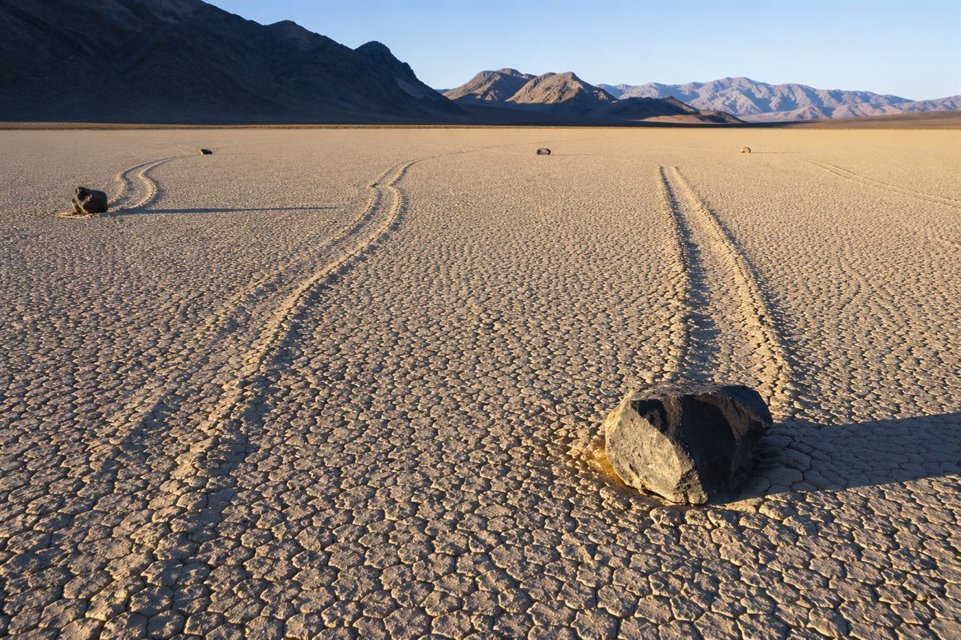 Several rocks each at the end of long furrow trails across the cracked mud floor of Racetrack Playa in Death Valley