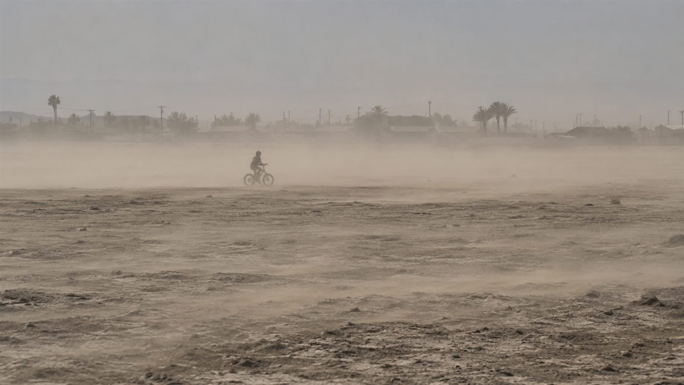 The white, salt-encrusted exposed lakebed of the Salton Sea stretching toward the water under a hazy sky, with dust visible above the flat surface