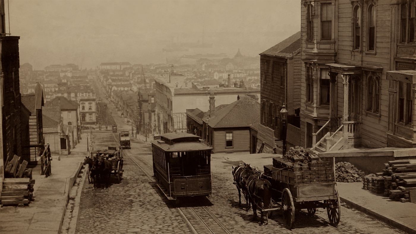 A vintage sepia view of a steep 1870s San Francisco street with an early cable car and horse-drawn wagons