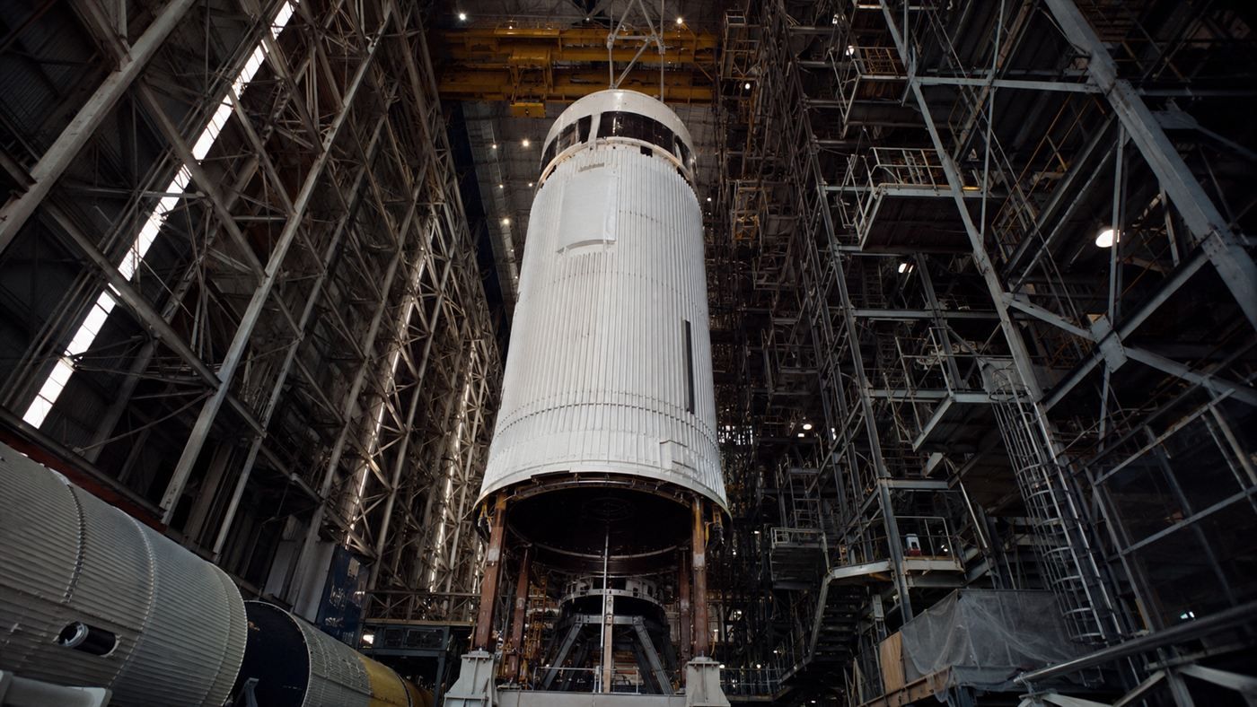 A towering rocket stage being assembled inside an enormous cavernous NASA-era assembly building