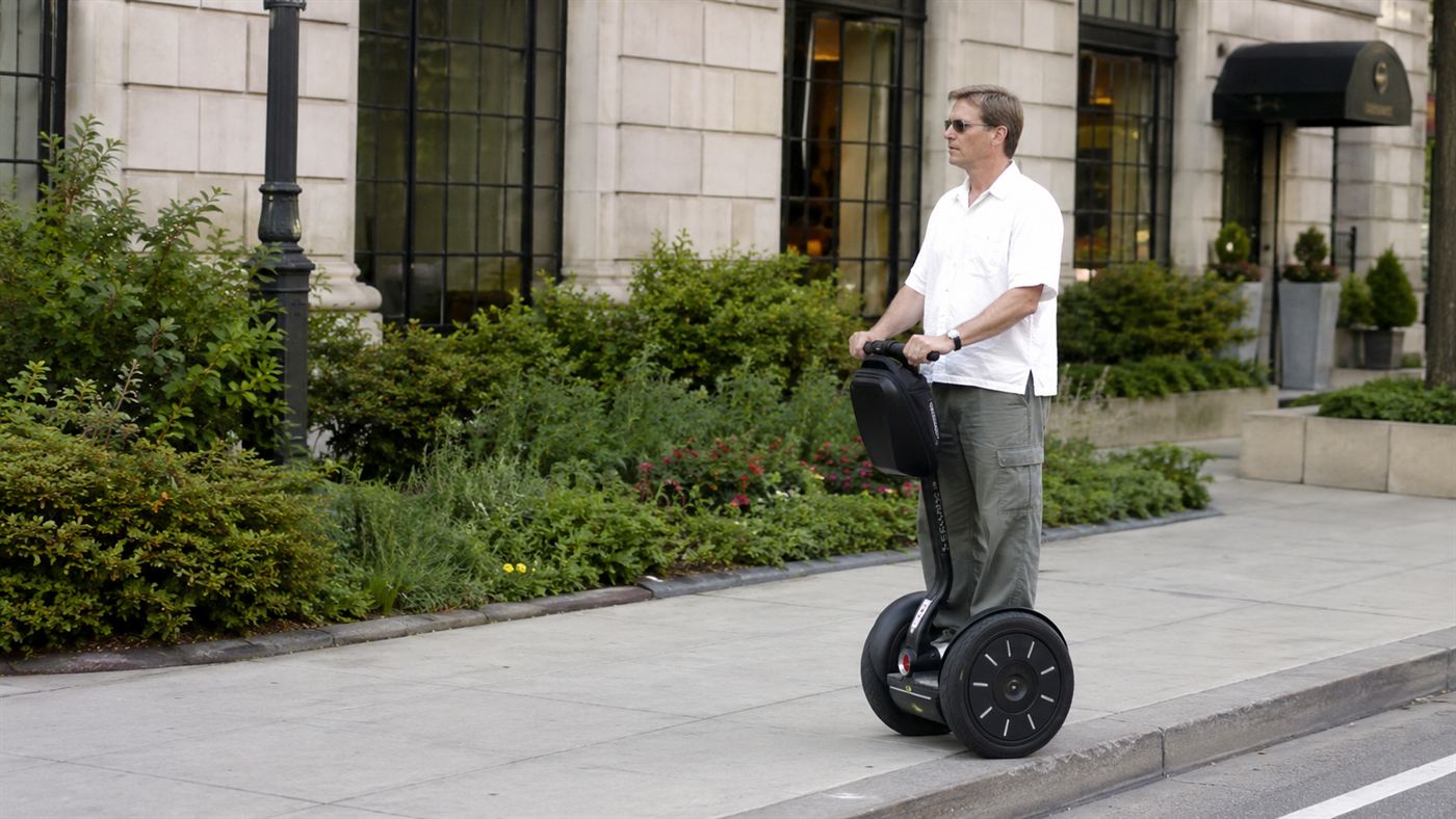 A person riding a two-wheeled self-balancing Segway personal transporter along a city pavement