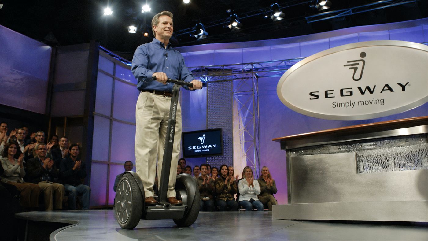 A 2001-style television studio reveal of a man riding a self-balancing two-wheeled personal transporter under bright lights