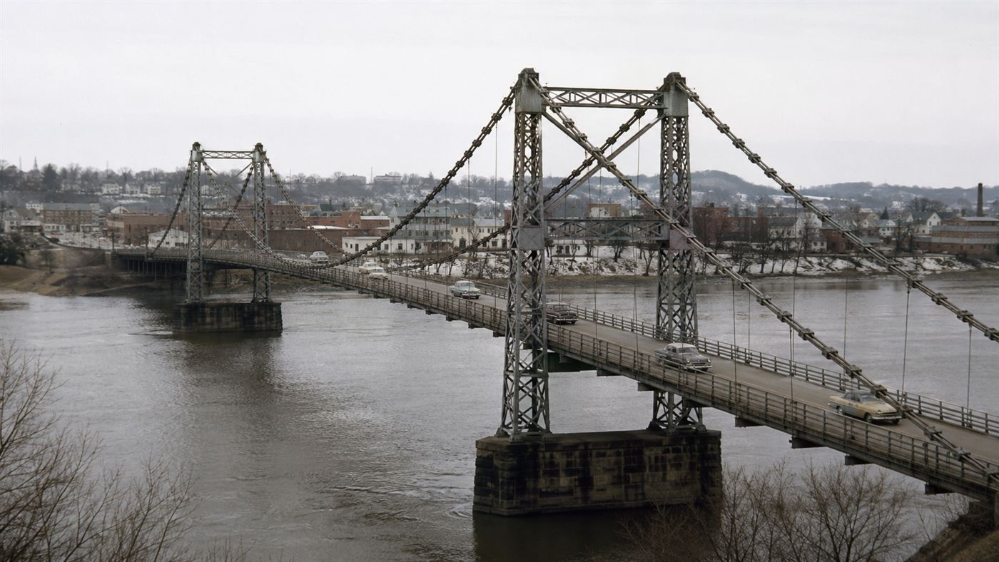 The old steel eyebar-chain Silver Bridge spanning a wide grey river between two towns under an overcast 1960s sky
