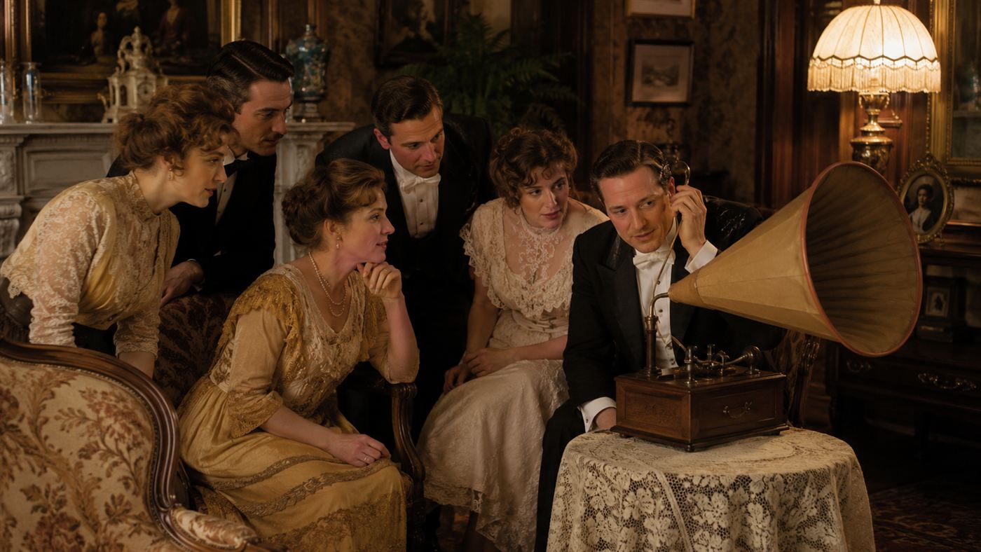 Well-dressed people in an early-1900s parlour listening to music from a telephone receiver fitted with a large paper funnel horn