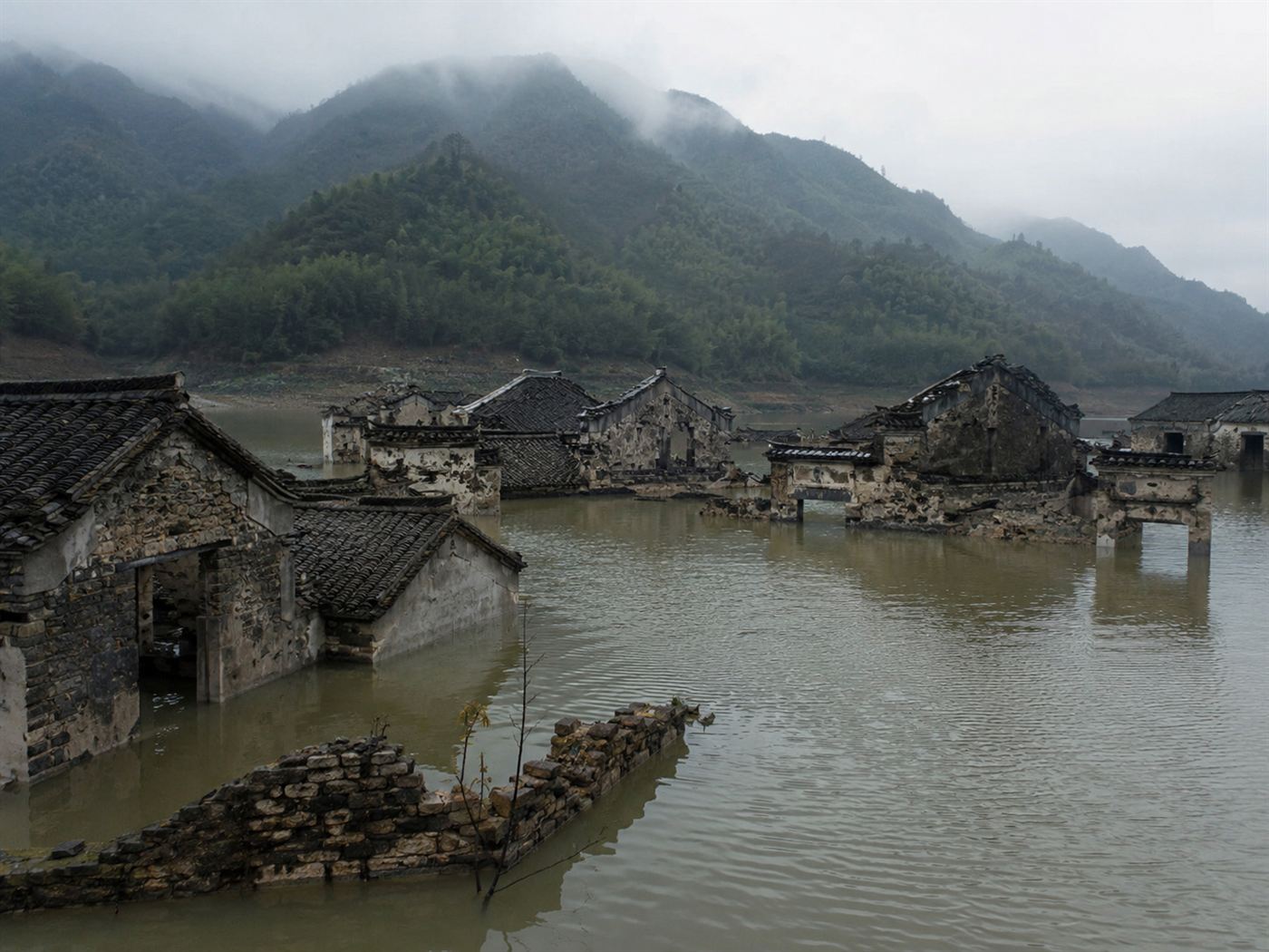 Partially submerged stone buildings of a flooded Chinese town rising above the brown water of the Three Gorges reservoir, ancient walls and rooflines emerging from the lake with misty mountains behind