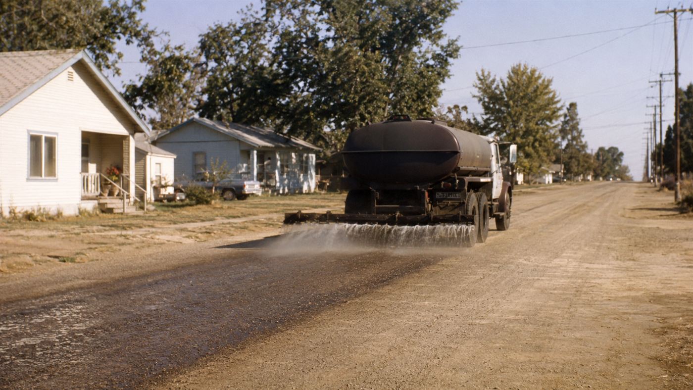 A 1970s tanker truck spraying oil onto a dusty dirt road in a rural American town