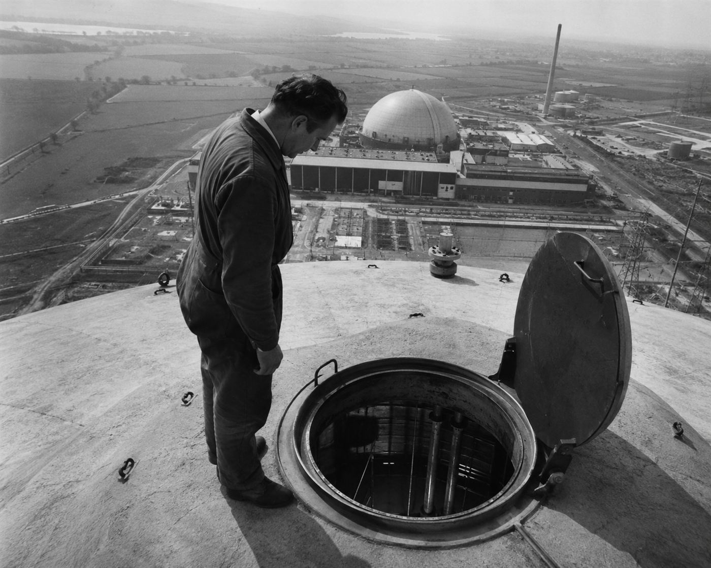 Industrial worker at the top of a large concrete nuclear reactor building looking down into an inspection hatch, high vantage point view of the Windscale facility structure, 1957