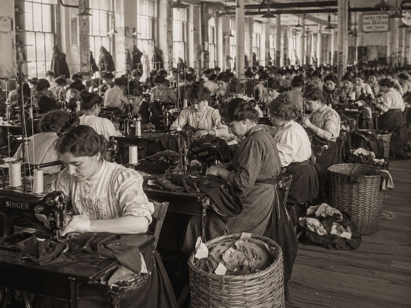 Young immigrant women working at sewing machines in a 1910s New York garment factory, rows of workers at crowded workstations with fabric and thread, early 20th century industrial labor conditions