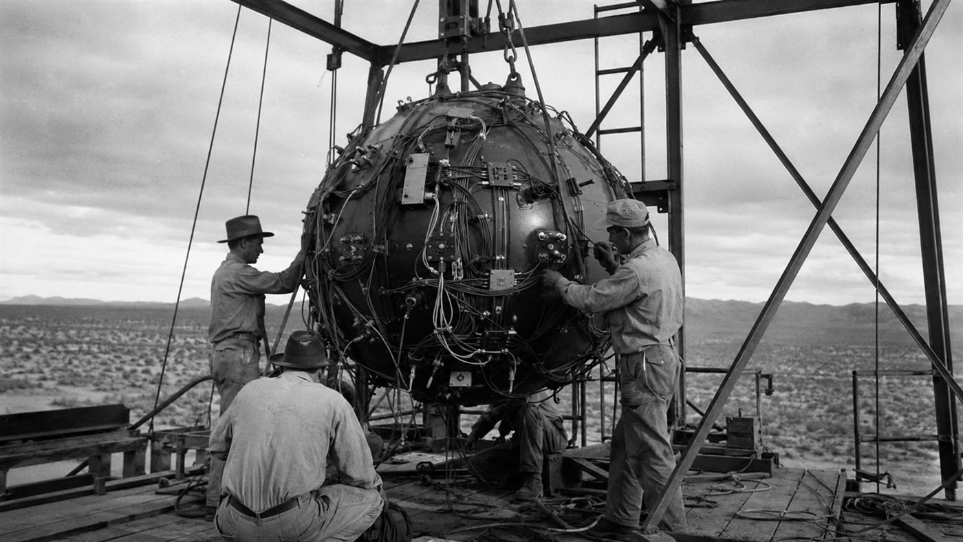 The Gadget, a large round metal atomic device, being assembled by technicians on a steel test tower in the New Mexico desert in 1945