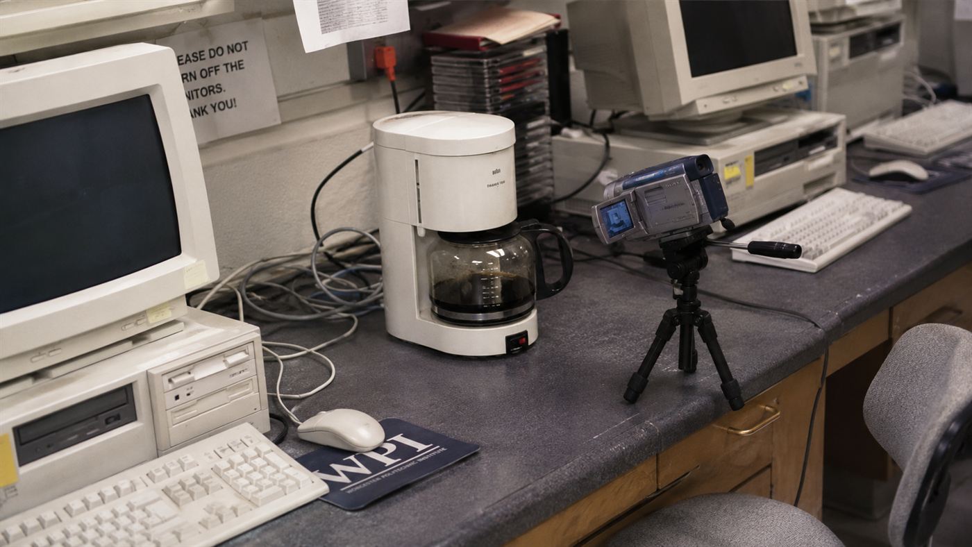 An old coffee machine in a 1990s computer lab with a small early video camera on a tripod pointed at it, the first webcam