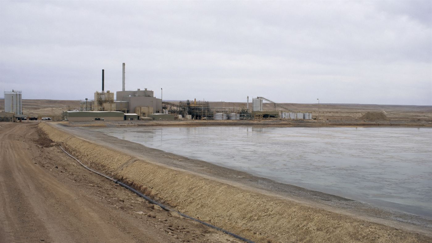 A 1970s uranium mill in the desert with a large earthen tailings waste pond and processing buildings under a pale sky