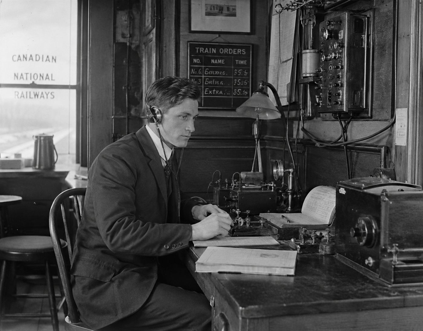 A railway telegraph dispatcher at his wooden desk with telegraph equipment in a small Canadian station office in 1917, determined expression, period uniform, black and white historical documentary photography