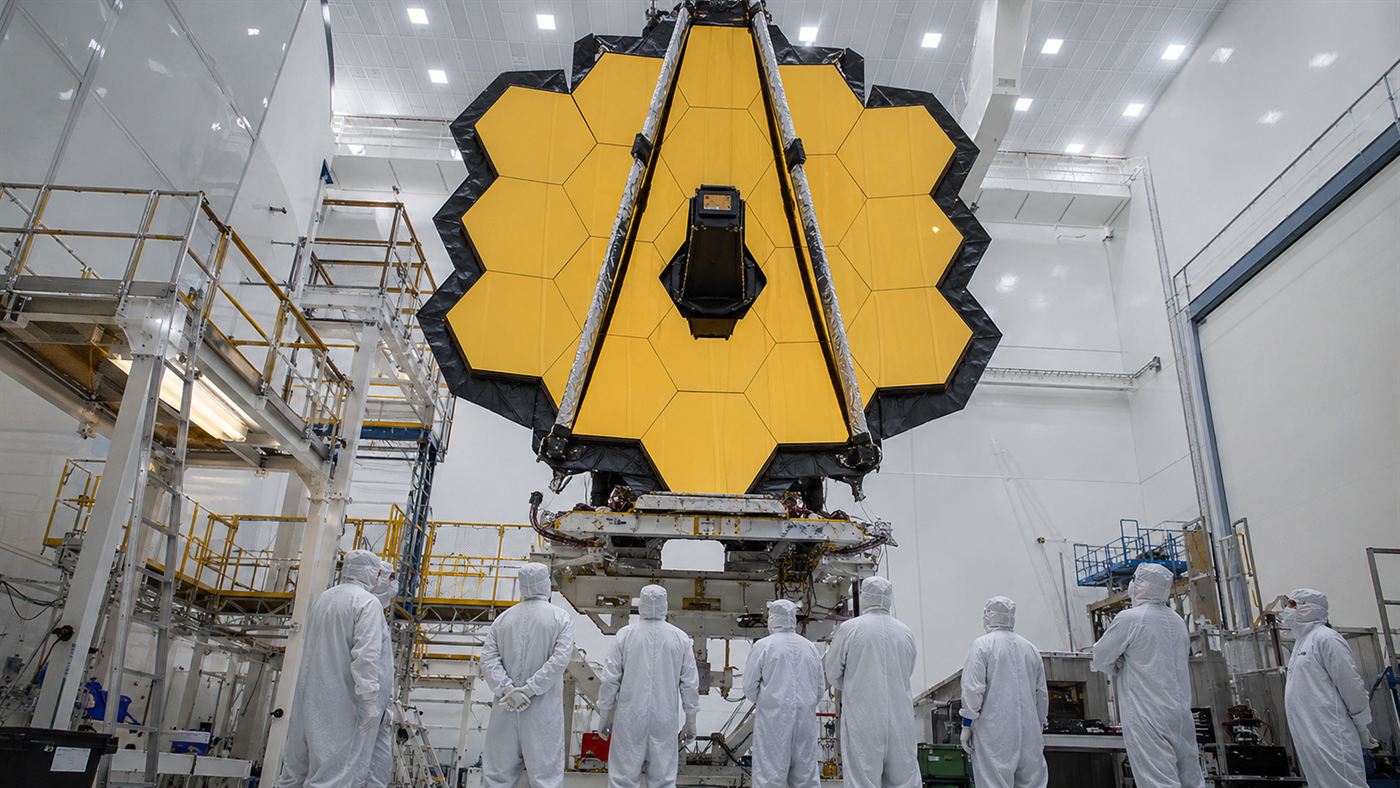 Engineers in white clean-room suits beneath the folded golden mirror of the James Webb Space Telescope