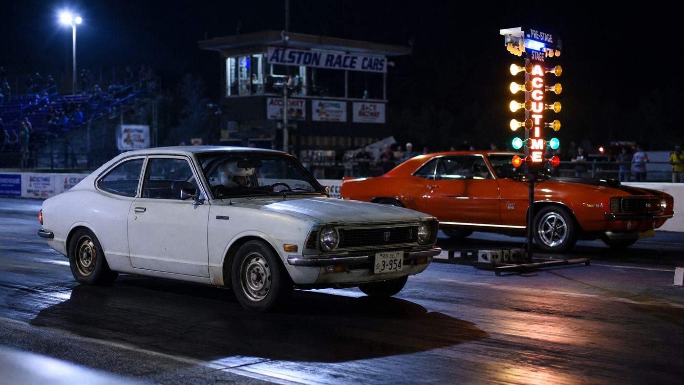 The White Zombie, a plain white 1972 Datsun electric drag car, staged at a drag strip start line beside a muscle car