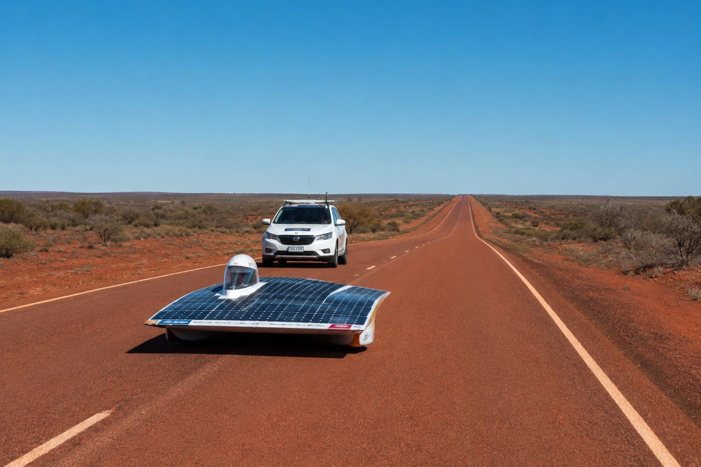A solar race car and its support vehicle on a long straight empty road through the red Australian desert under a clear sky