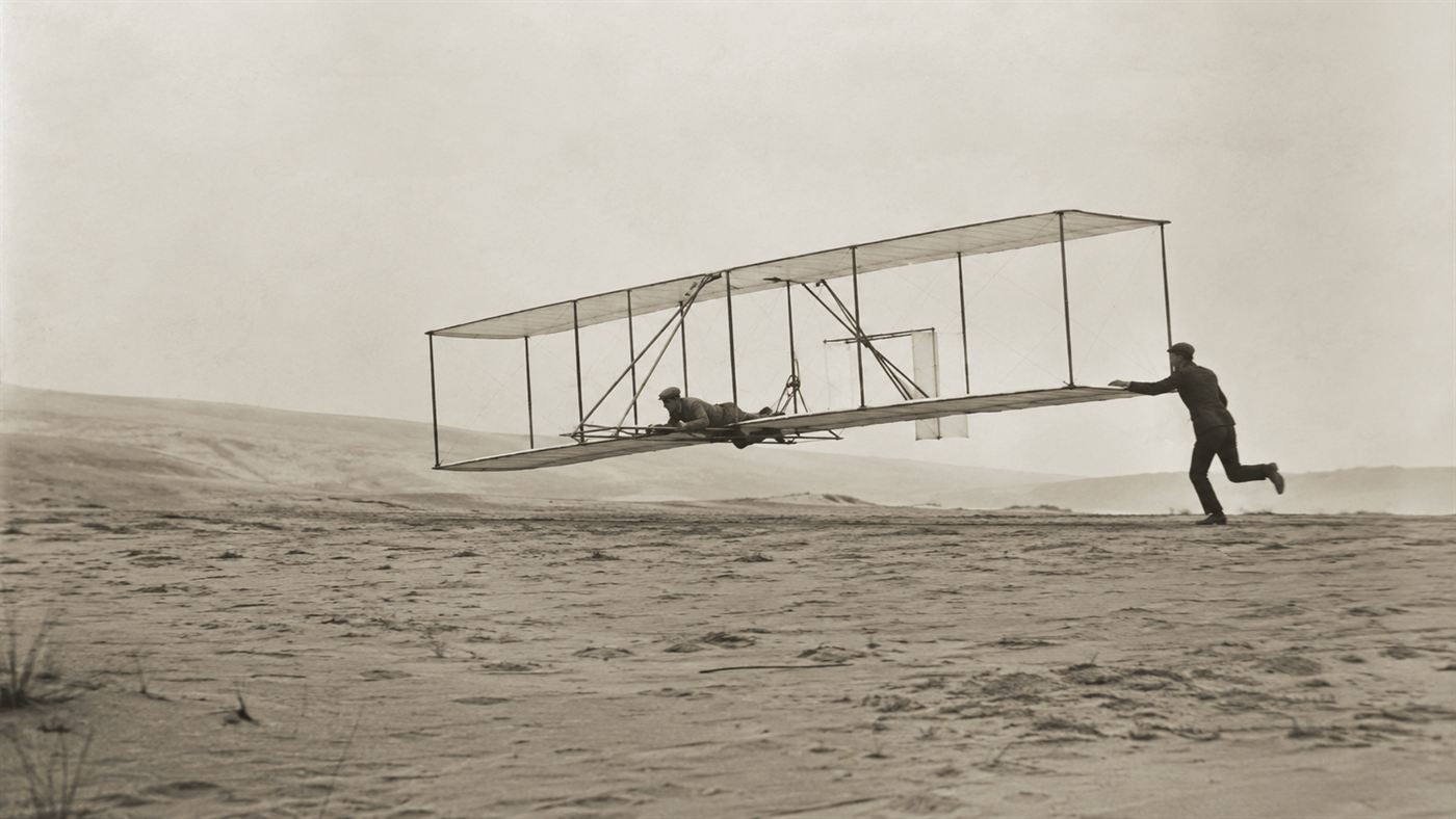 The Wright brothers' fragile 1903 Flyer lifting off the sand at Kitty Hawk, one brother at the controls and the other running alongside
