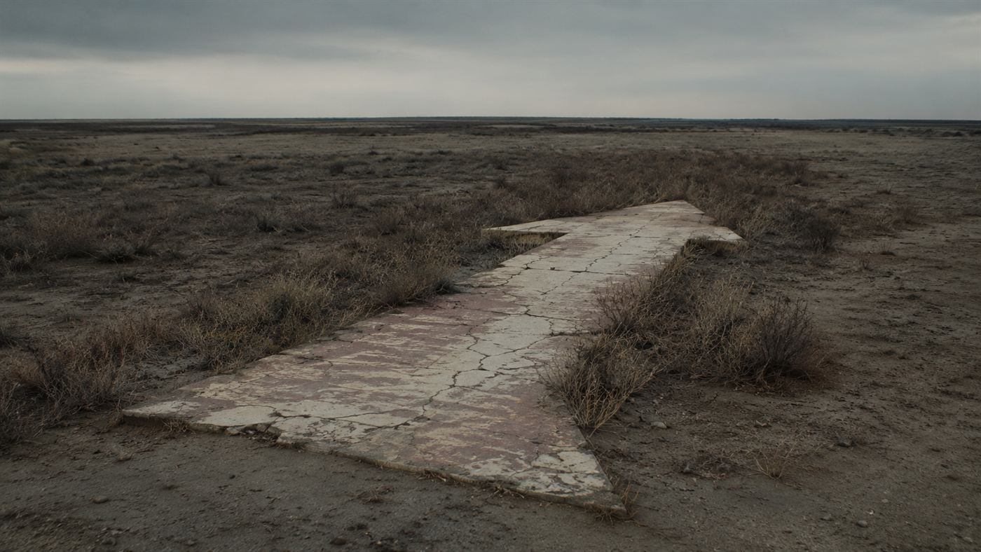 A cracked and weathered concrete arrow half overgrown with scrub in an empty landscape, its beacon tower long gone
