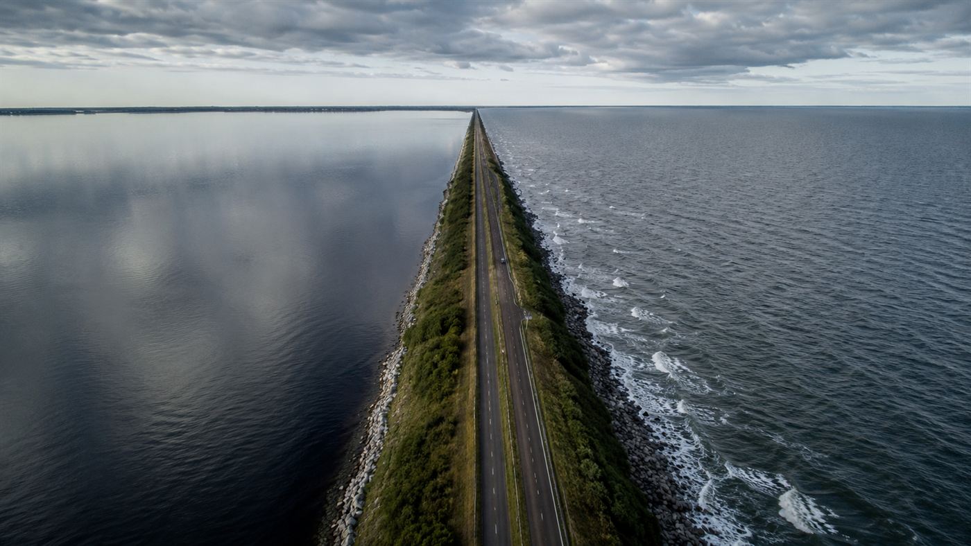 A long straight causeway dike stretching to the horizon, separating a calm freshwater lake from the open grey sea