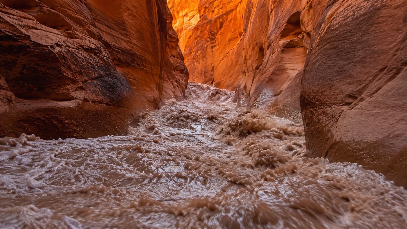 Muddy brown flash flood water surging through a narrow slot canyon in the Arizona desert, filling the tight sandstone passage