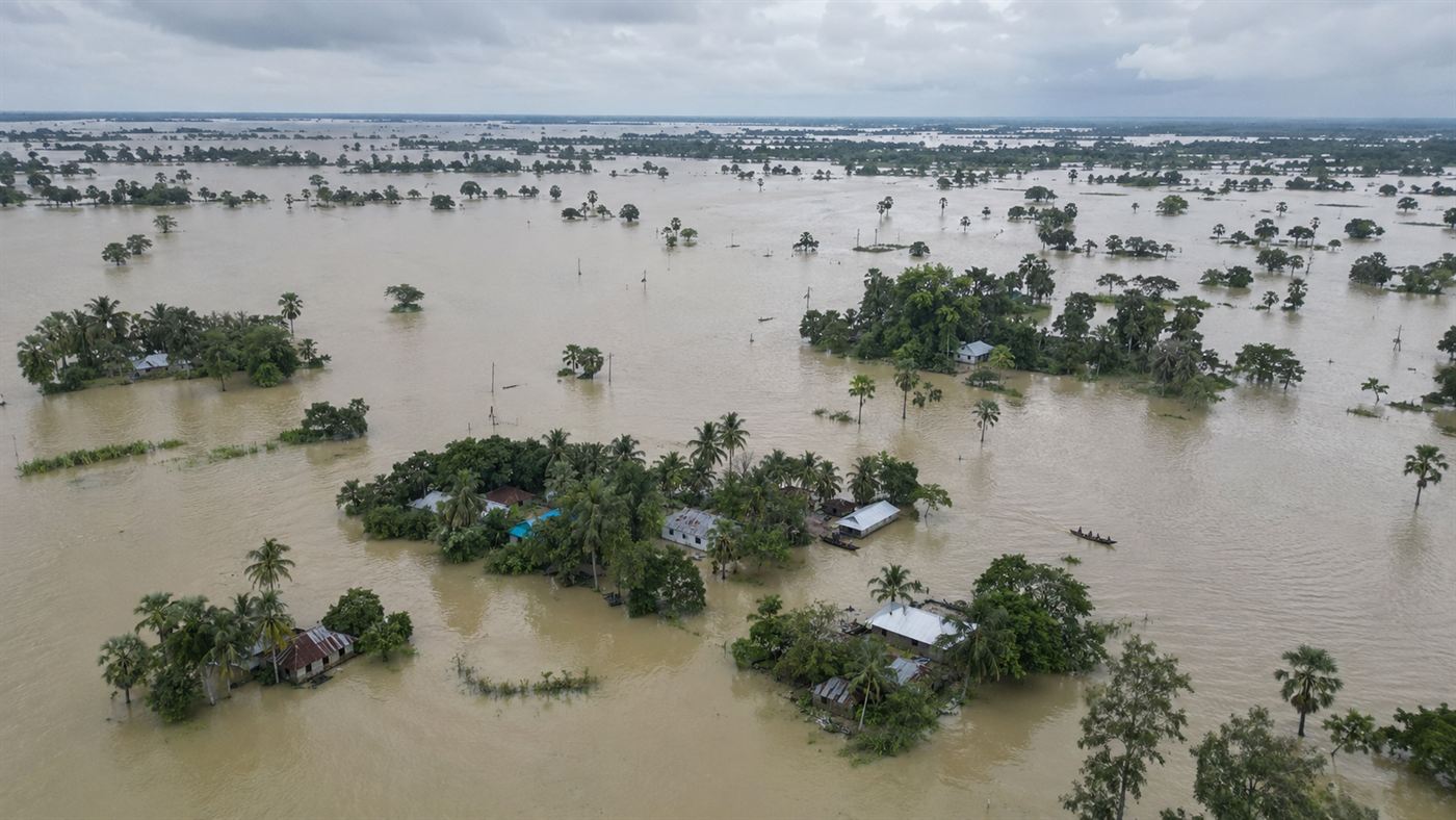 An aerial view of a flooded monsoon landscape in Bangladesh, villages and rice fields half submerged under brown water with houses and palm trees standing above it and small boats moving between them