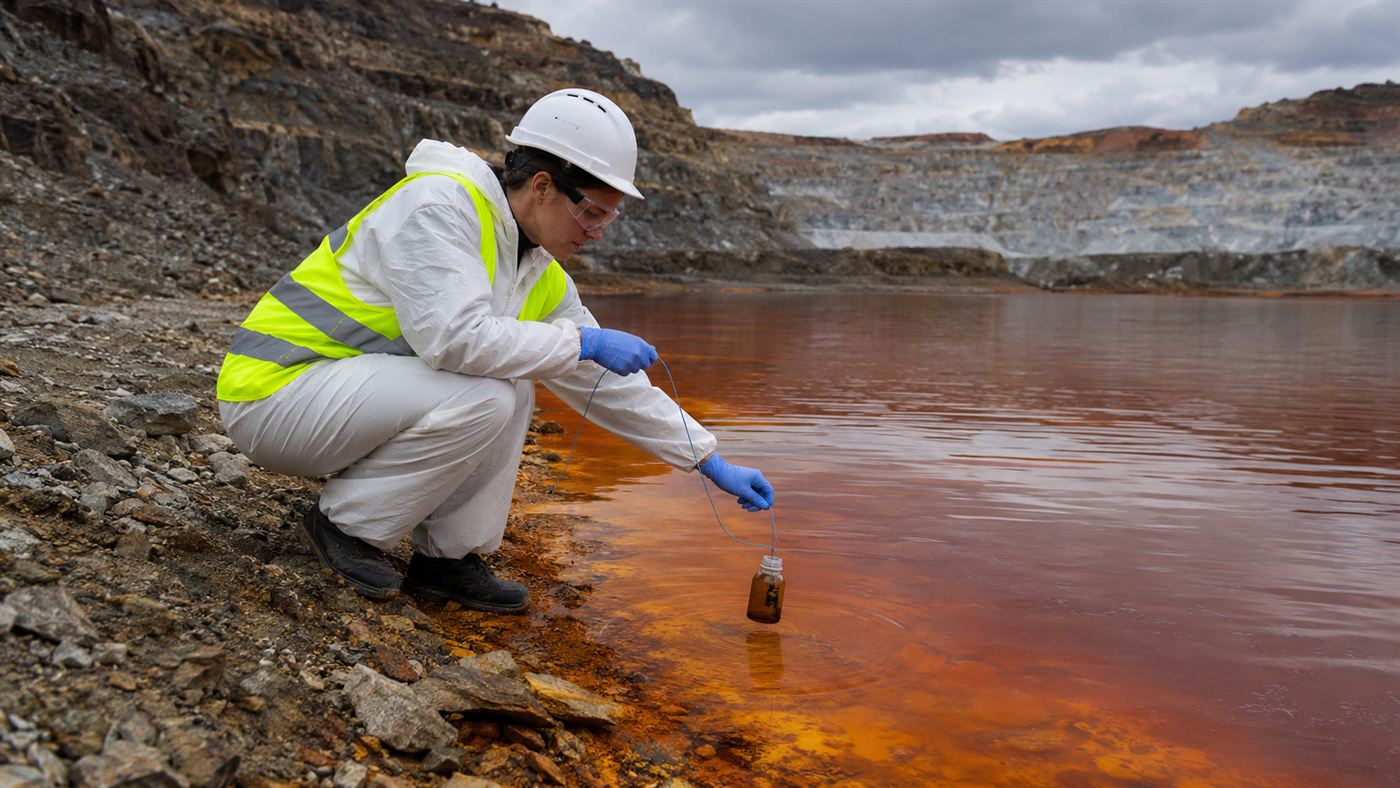 A researcher in gloves collecting a sample of acidic water at the edge of the Berkeley Pit to study its extremophiles