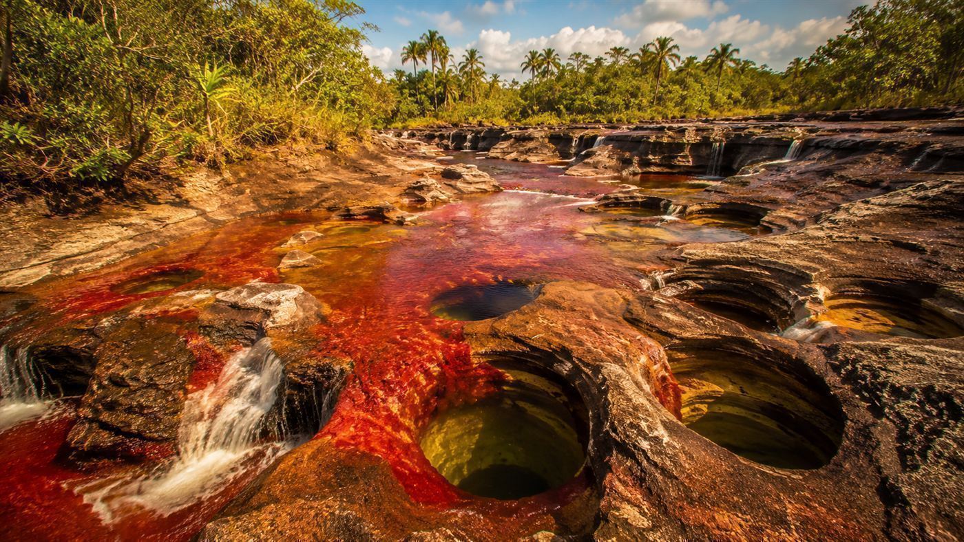 The colourful river flowing through ancient rock formations with small waterfalls and round potholes carved into the stone